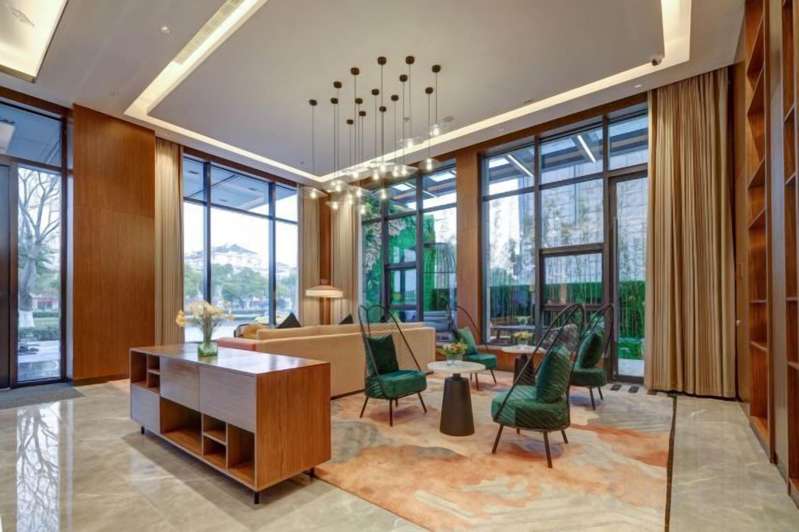 A modern interior with a wooden reception desk, green chairs, a sofa, and large windows offering a view of greenery and buildings.