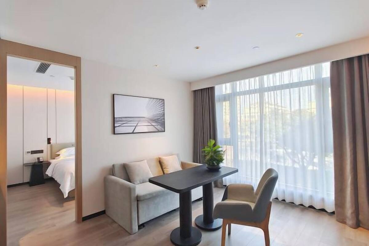 Una habitación de hotel moderna con cama, sofá, mesa de comedor y un ventanal con cortinas.