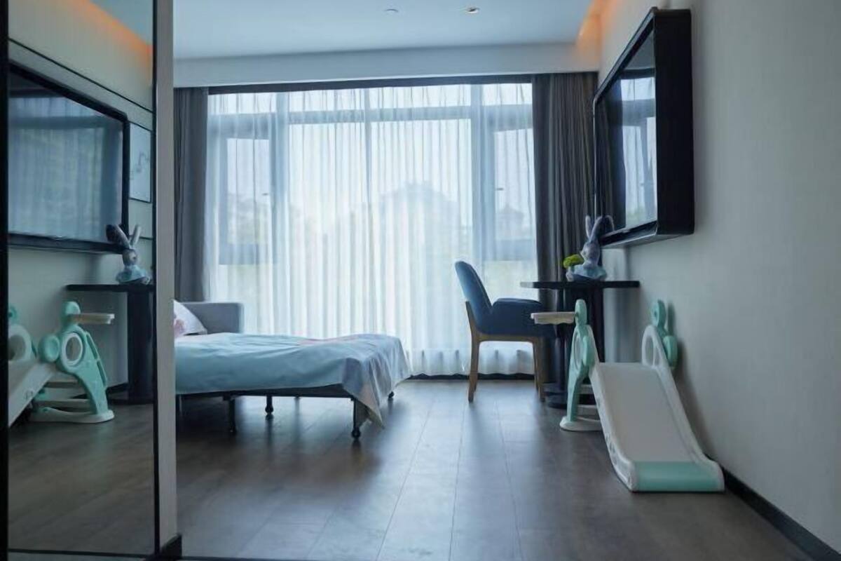 Una habitación de hotel con cama, una silla, un escritorio, un televisor y un ventanal con cortinas.