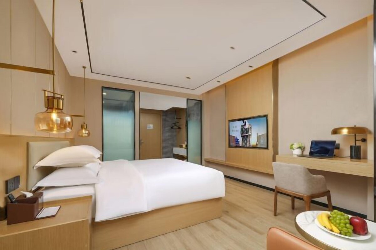 A modern hotel room with a bed, desk, chair, and a dining area with fruit.