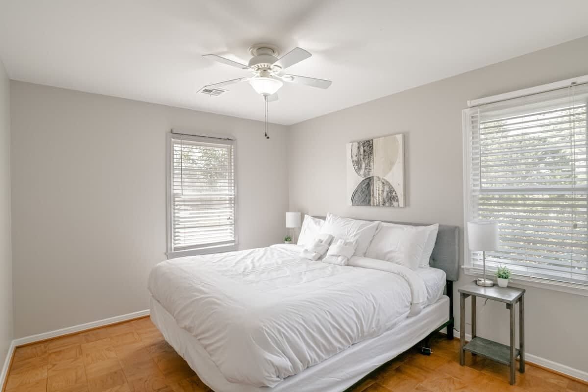 A bedroom with a bed, a ceiling fan, a nightstand with a lamp, and a window with blinds.