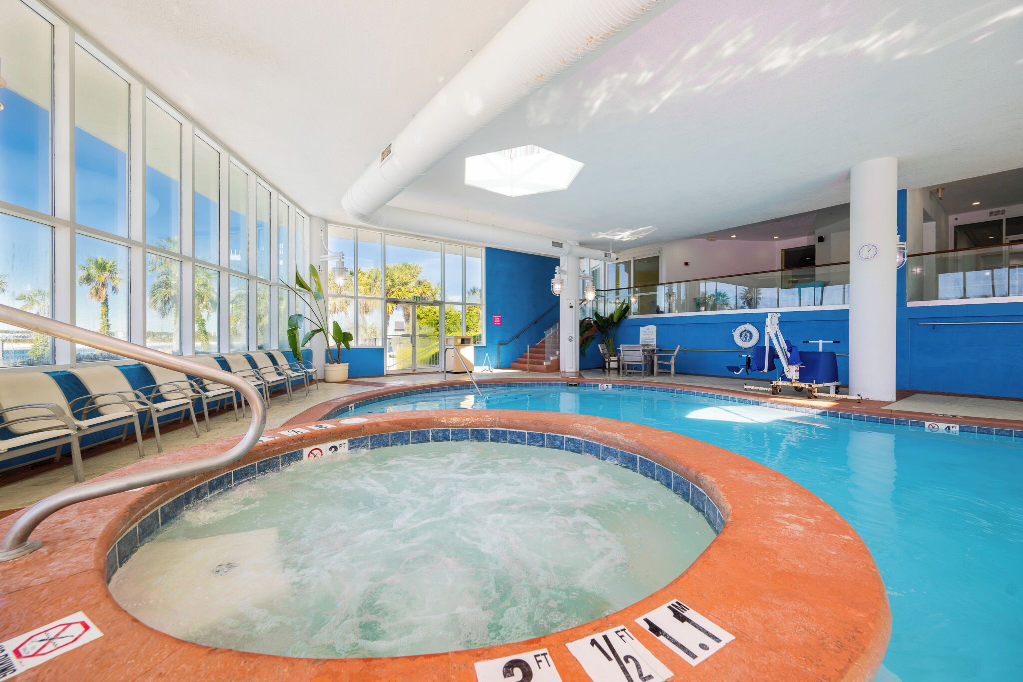 An indoor swimming pool with a hot tub, seating area, and a view of palm trees outside.