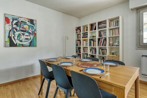 Ein Essbereich mit einem Holztisch, gedeckt für vier Personen, umgeben von Stühlen. Eine Bücherwand mit Büchern und Dekoration befindet sich zur Rechten.