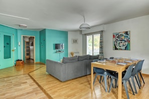 Ein modernes Wohnzimmer mit grauem Sofa, Holzesstisch und blauen Stühlen. Ein Fernseher steht auf einem Regal, ein Gemälde hängt an der Wand und ein Fenster ist mit Vorhängen drapiert.