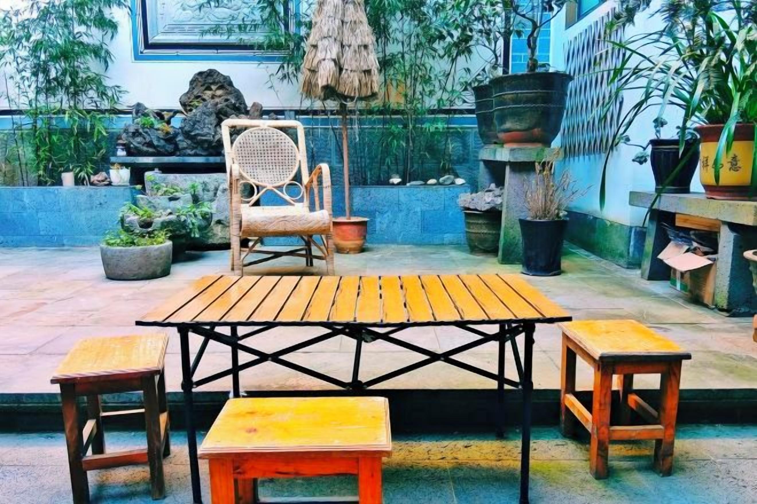 Un patio al aire libre con una mesa y sillas de madera, rodeado de macetas con plantas y un banco de piedra.