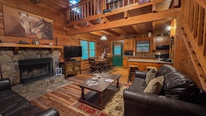 A cozy cabin interior with a stone fireplace, leather sofa, wooden dining table, and a kitchen area. - Labor Day at Pike's Peak - Wifi and Hot Tub! - Callaway Gardens (Warm Springs)