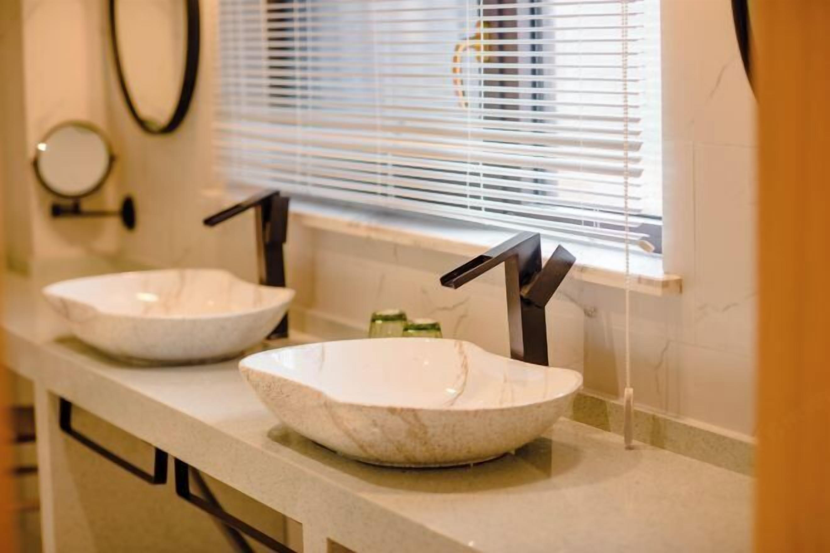 A bathroom with two white marble sinks, black faucets, and a window with blinds.