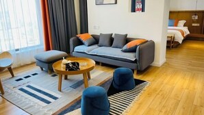 Un saló modern amb un sofà, un reposapius i una taula de centre. Hi ha un llit al fons i una finestra amb cortines.