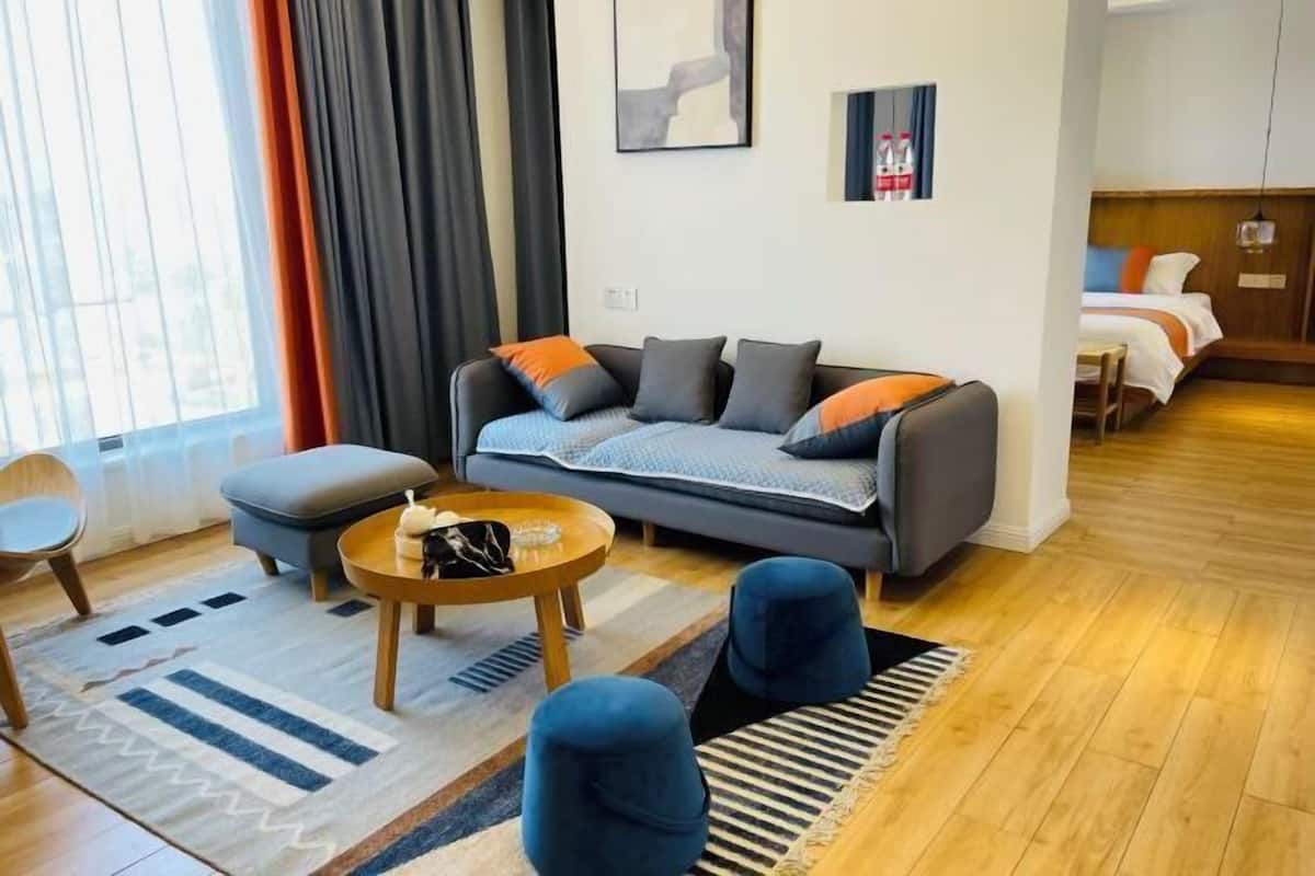 A modern living room with a sofa, ottoman, and coffee table. There is a bed in the background and a window with curtains.