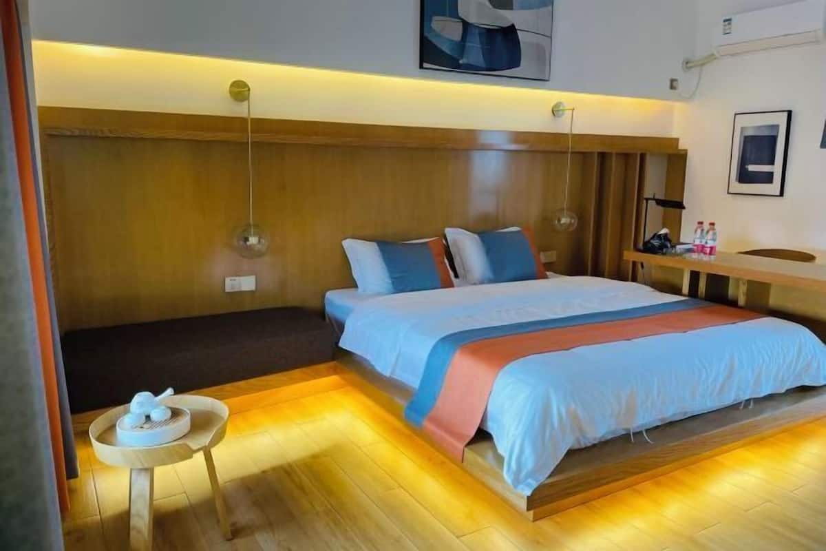 A modern hotel room with a wooden headboard, a large bed with blue and orange bedding, a small round table with a tea set, and a bedside table with bottles.