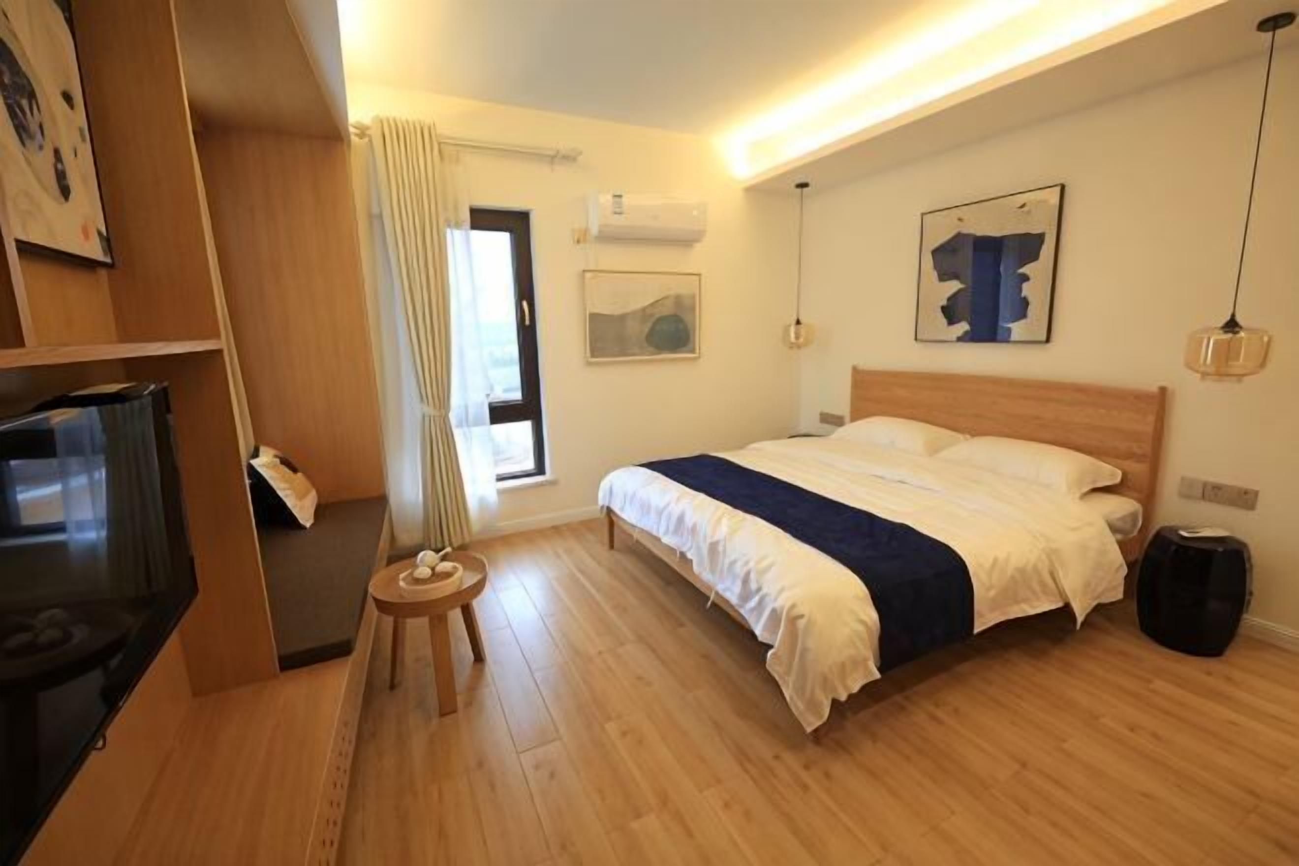 A hotel room with a bed, a TV, a small table, a chair, a window with curtains, and artwork on the walls.
