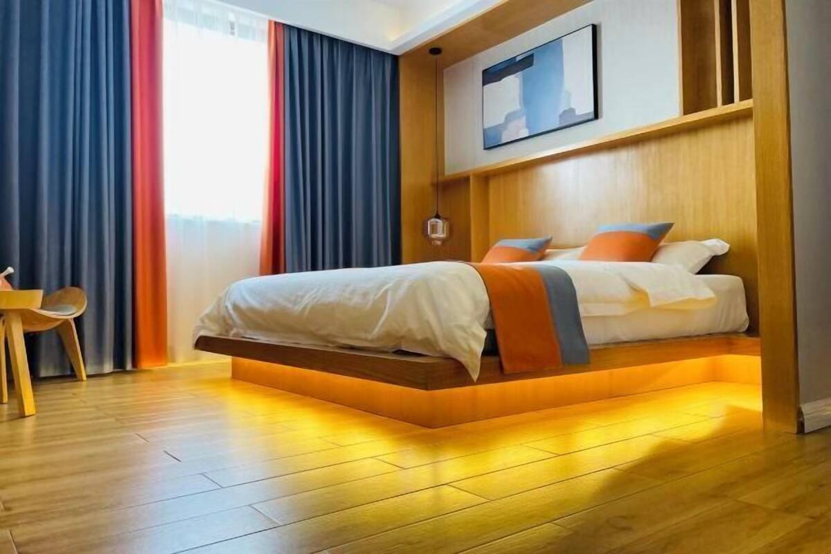 A modern hotel room with a bed, wooden flooring, and a view of the city through large windows.