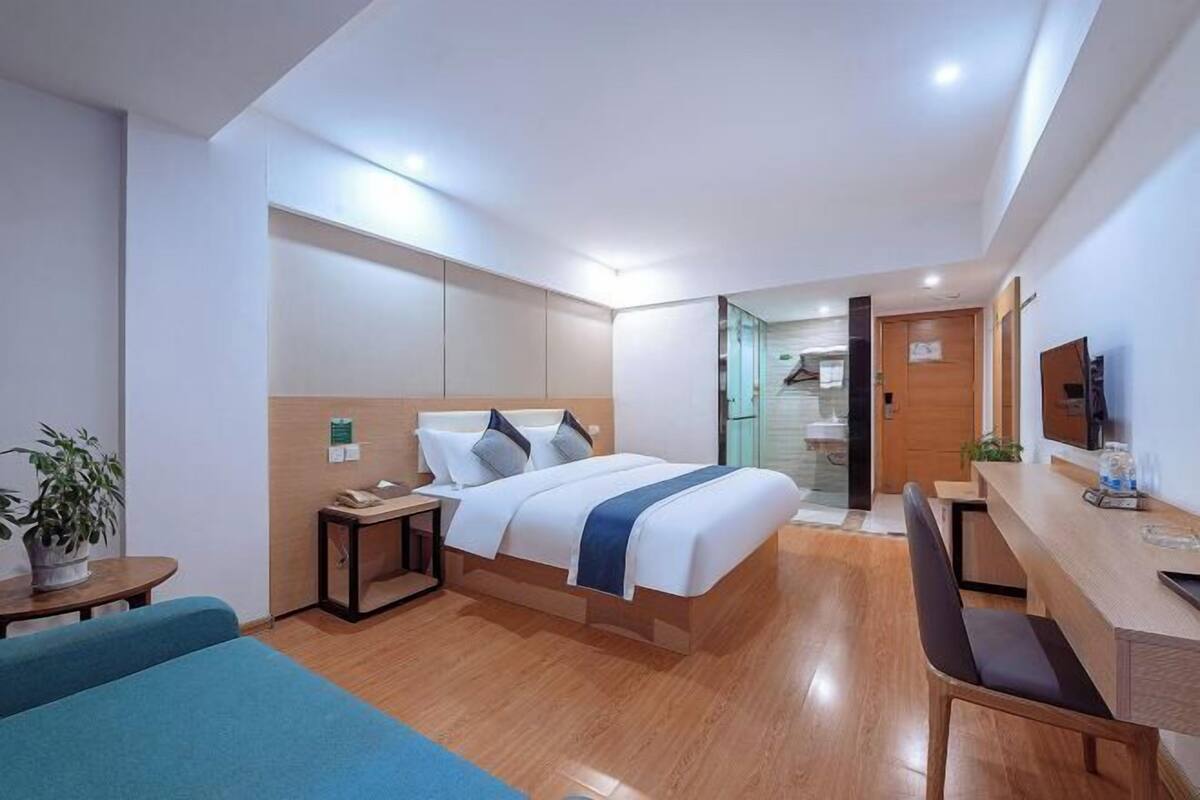 A modern hotel room with a bed, bedside table, desk, chair, and a potted plant.