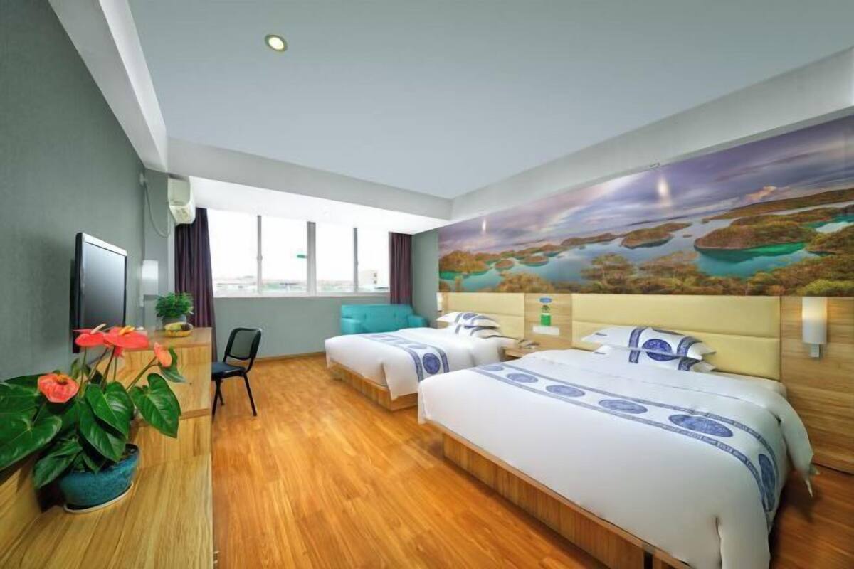 A hotel room with two beds, a large window, a painting, and a plant.