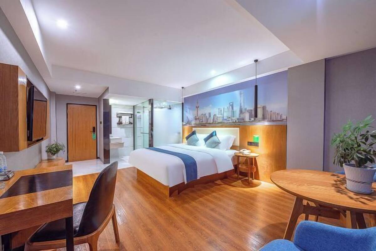 A modern hotel room with a wooden floor, a bed with a blue accent, a desk, and a dining area.