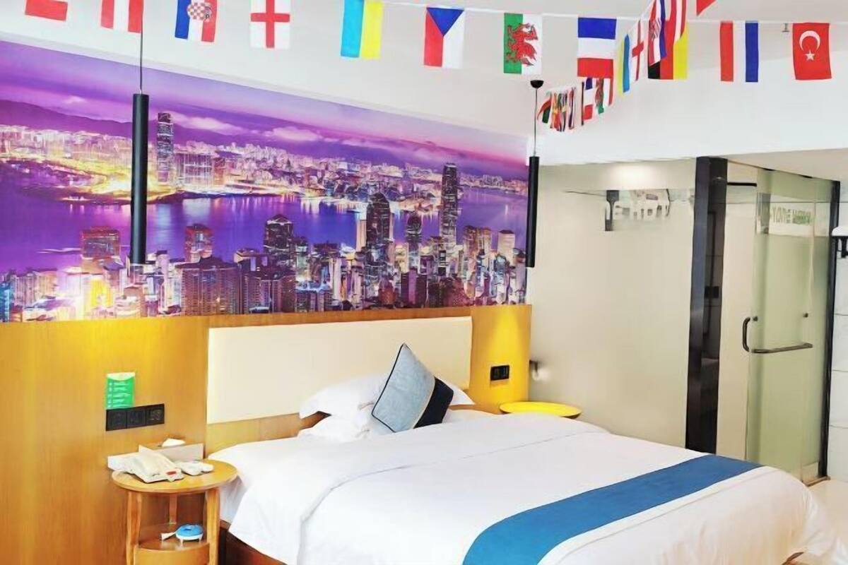 A hotel room with a cityscape view, a bed with white bedding, a bedside table with a lamp, and a digital clock.