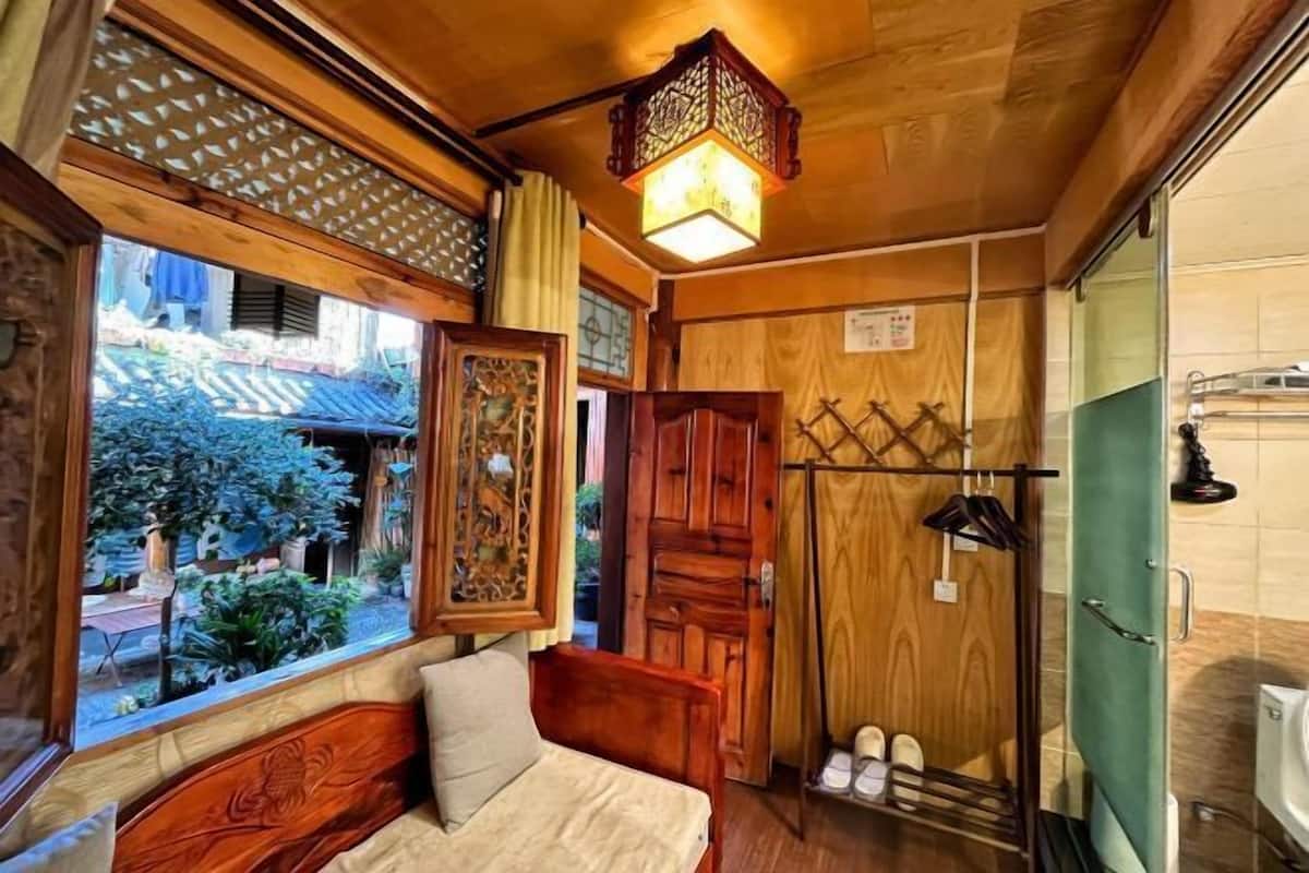A wooden room with a bed, a window with a view of greenery, a wooden door, and a shoe rack.