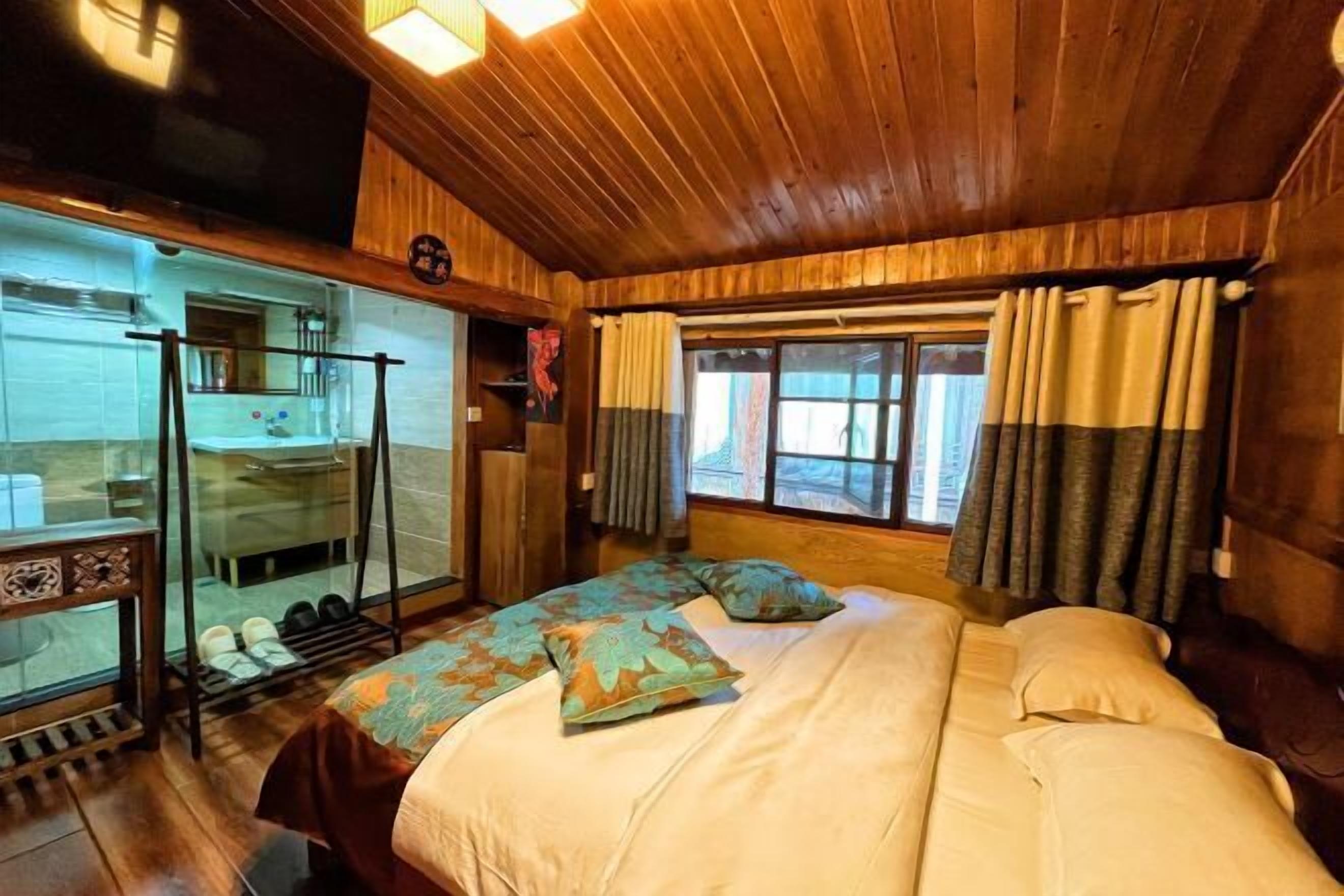 A wooden cabin room with a bed, a glass shower enclosure, and a window with curtains.