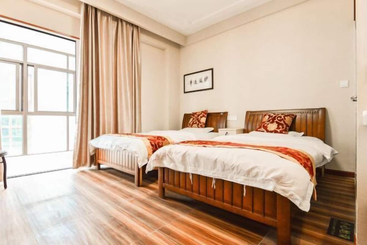 A hotel room with two wooden beds, a large window with curtains, a framed picture on the wall, and a wooden floor.