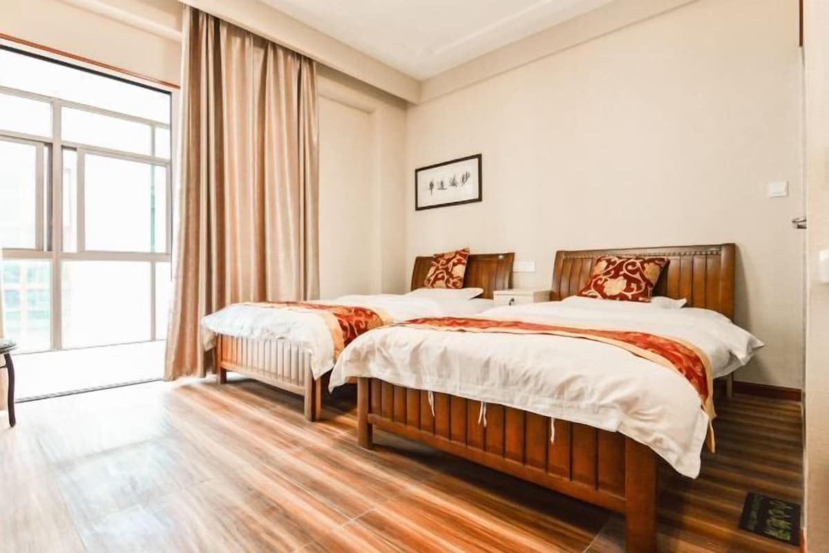 A hotel room with two wooden beds, a large window with curtains, and a framed picture on the wall.