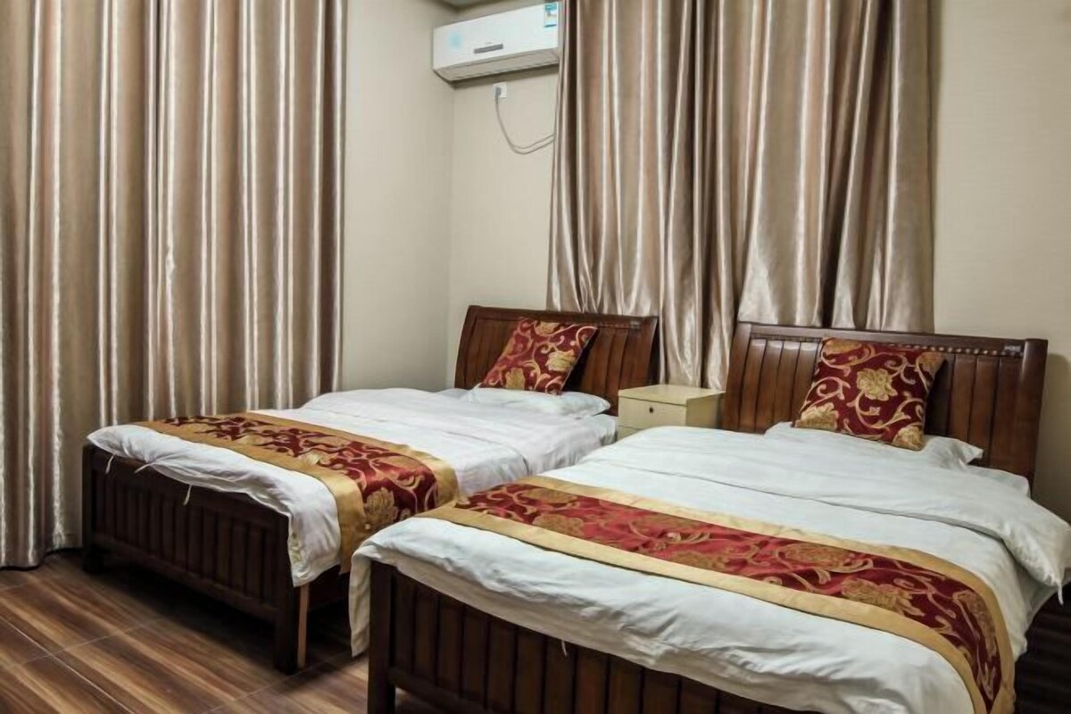 A hotel room with two wooden beds, beige curtains, and a white air conditioning unit.