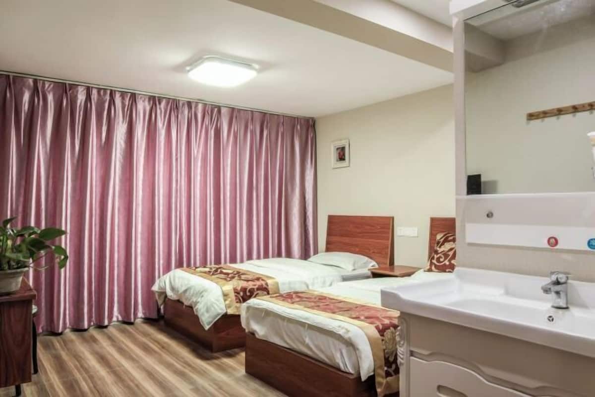 A hotel room with two beds, a large window with curtains, a wooden headboard, a white sink, and a potted plant.