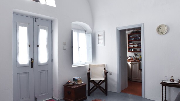 A hallway with white walls, a wooden chair, a small table with books, and a kitchen visible through an open door. - Calderimi,Traditional house in the center of Santorini. (FIRA)