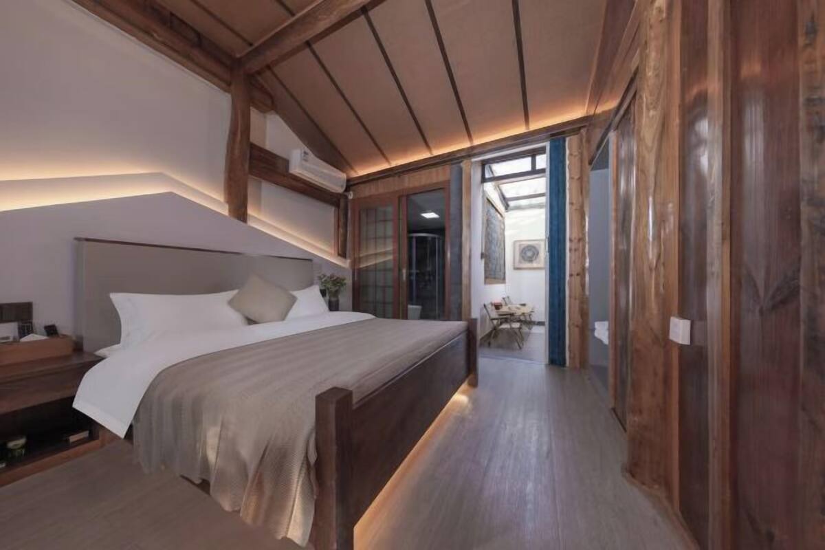Une chambre avec un lit en bois, une table de chevet et une commode. Un couloir mène à une salle à manger.