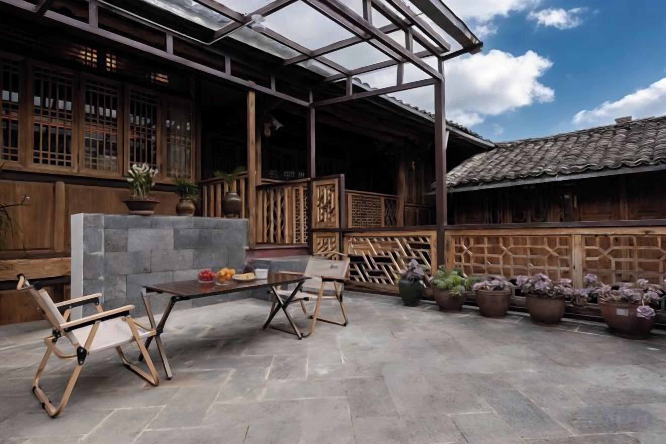 Un edificio de madera tradicional con un patio cubierto que tiene una mesa de piedra, dos sillas de madera y plantas en macetas.
