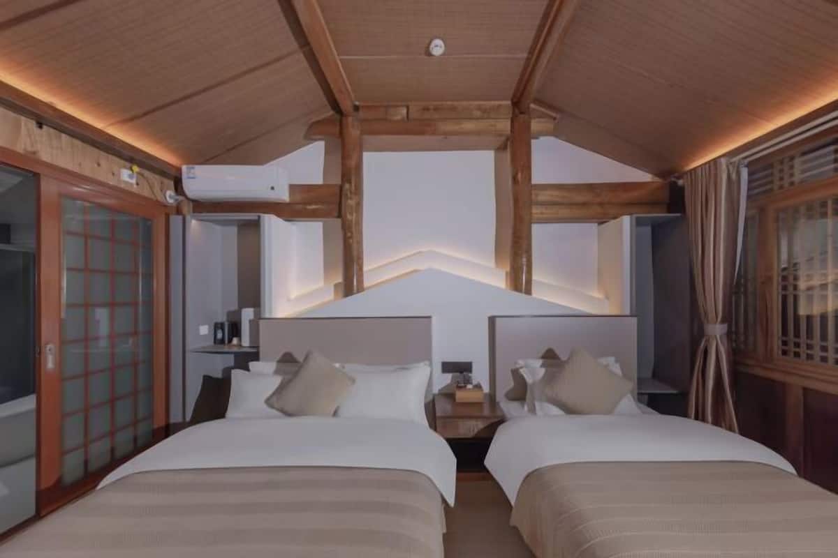 Une chambre d’hôtel avec deux lits, des poutres apparentes en bois au plafond, un luminaire moderne et une porte-fenêtre coulissante.