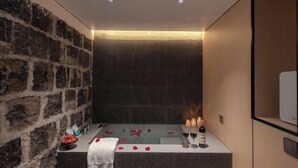 Um quarto de spa com parede de pedra, uma banheira com pétalas de rosa, velas e uma garrafa de vinho.