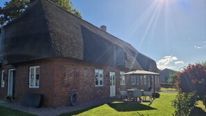 A brick house with a thatched roof, a patio with outdoor furniture, and a well-maintained lawn. - Vacation apartment Friesenwind (Rantrum)
