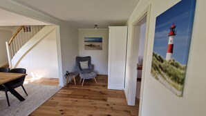 A room with a wooden floor, a dining table with chairs, a grey armchair, a small side table, and a painting of a lighthouse. - Vacation apartment Friesenwind (Rantrum)