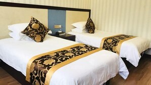 Dos camas individuales con ropa blanca y bordes dorados decorativos, una mesita de noche con lámpara, y una pared a rayas en una habitación de hotel.