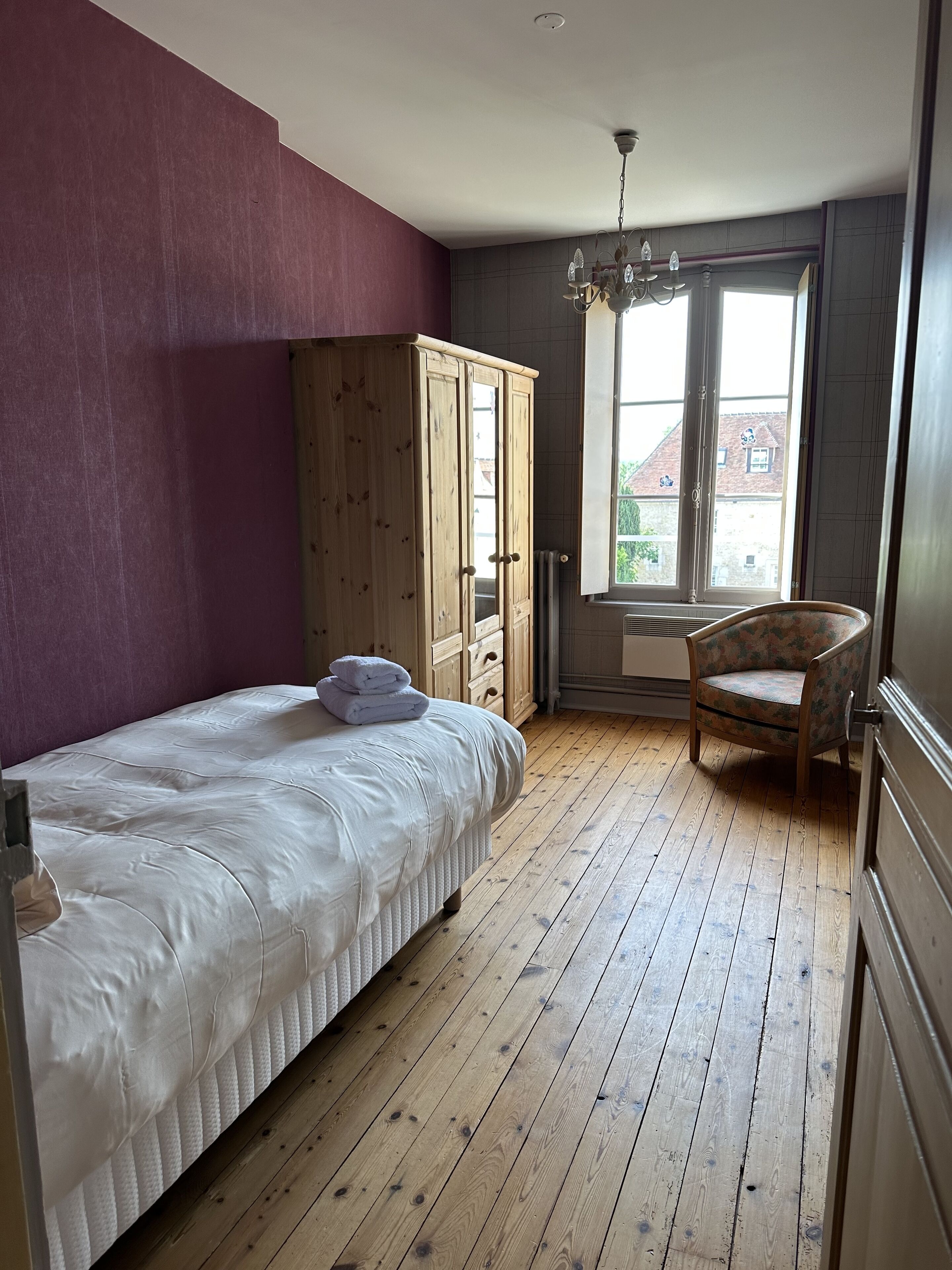 Een slaapkamer met een bed, een houten kledingkast, een stoel en een raam met uitzicht op gebouwen.