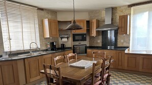Una cocina con armarios de madera, una mesa de comedor con sillas, un horno empotrado y un ventanal con persianas.