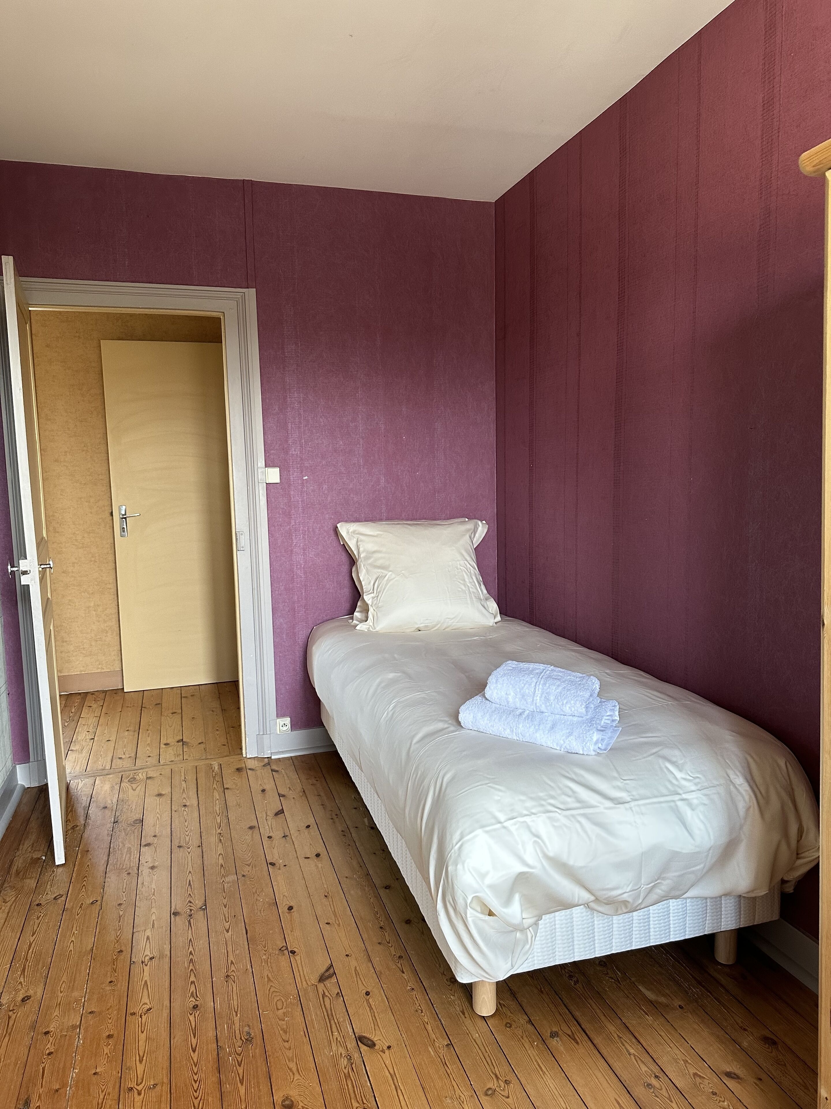 Ein Einzelbett mit weißer Matratze und Kissen, Holzboden und eine Tür zu einem anderen Raum.