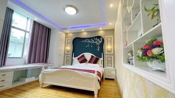 A bedroom with a bed, bedside tables, a desk, a window with curtains, and a wall-mounted shelf with flowers.
