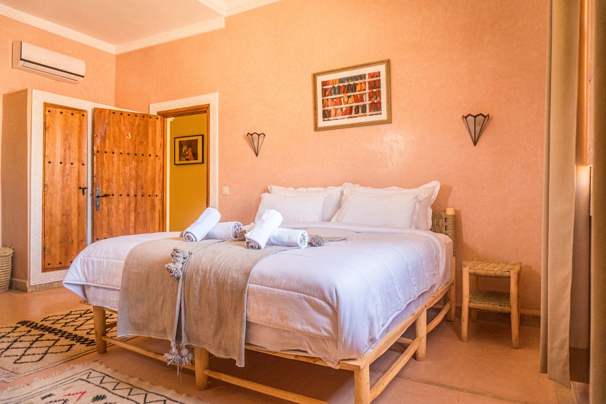 A double bed with a beige blanket, a small wooden stool, and a framed picture on the wall.