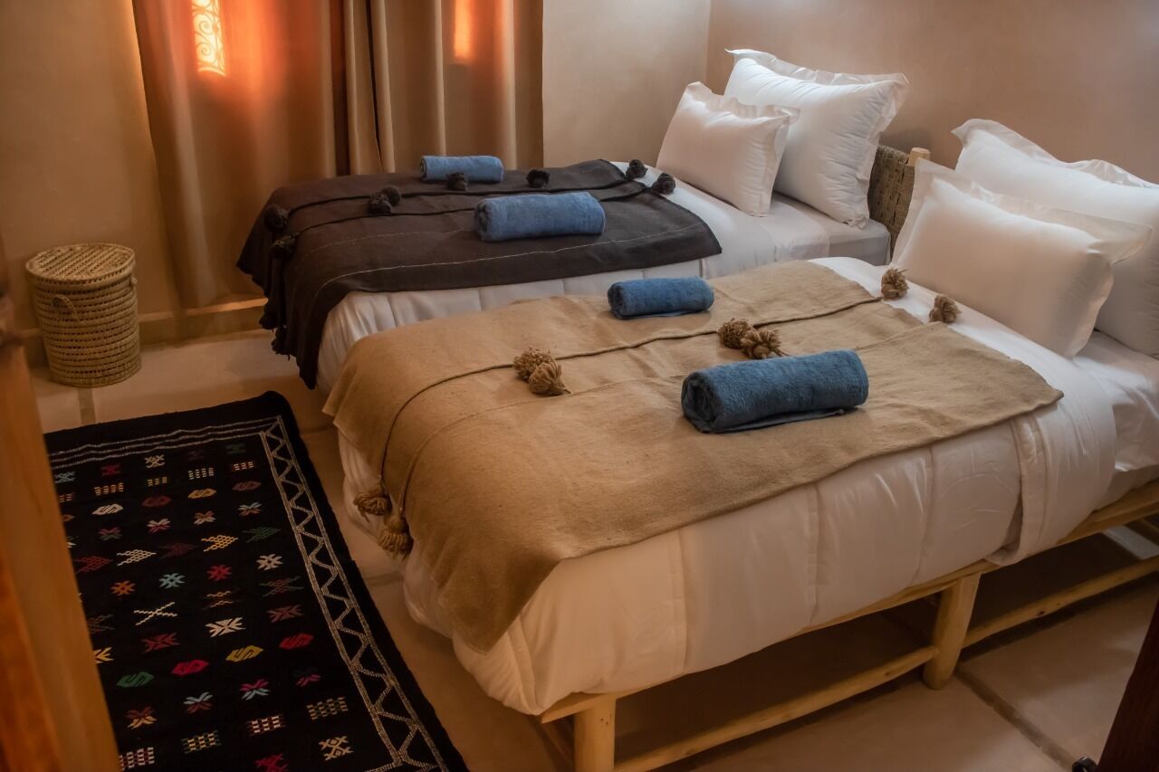 Ein Hotelzimmer mit zwei Betten, jedes mit einer Decke und Kissen in unterschiedlicher Farbe. Auf dem Boden steht ein gewebter Korb und ein gemustertes Teppich.