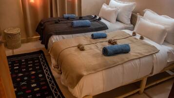 Ein Hotelzimmer mit zwei Betten, jedes mit einer Decke und Kissen in unterschiedlicher Farbe. Auf dem Boden steht ein gewebter Korb und ein gemustertes Teppich.