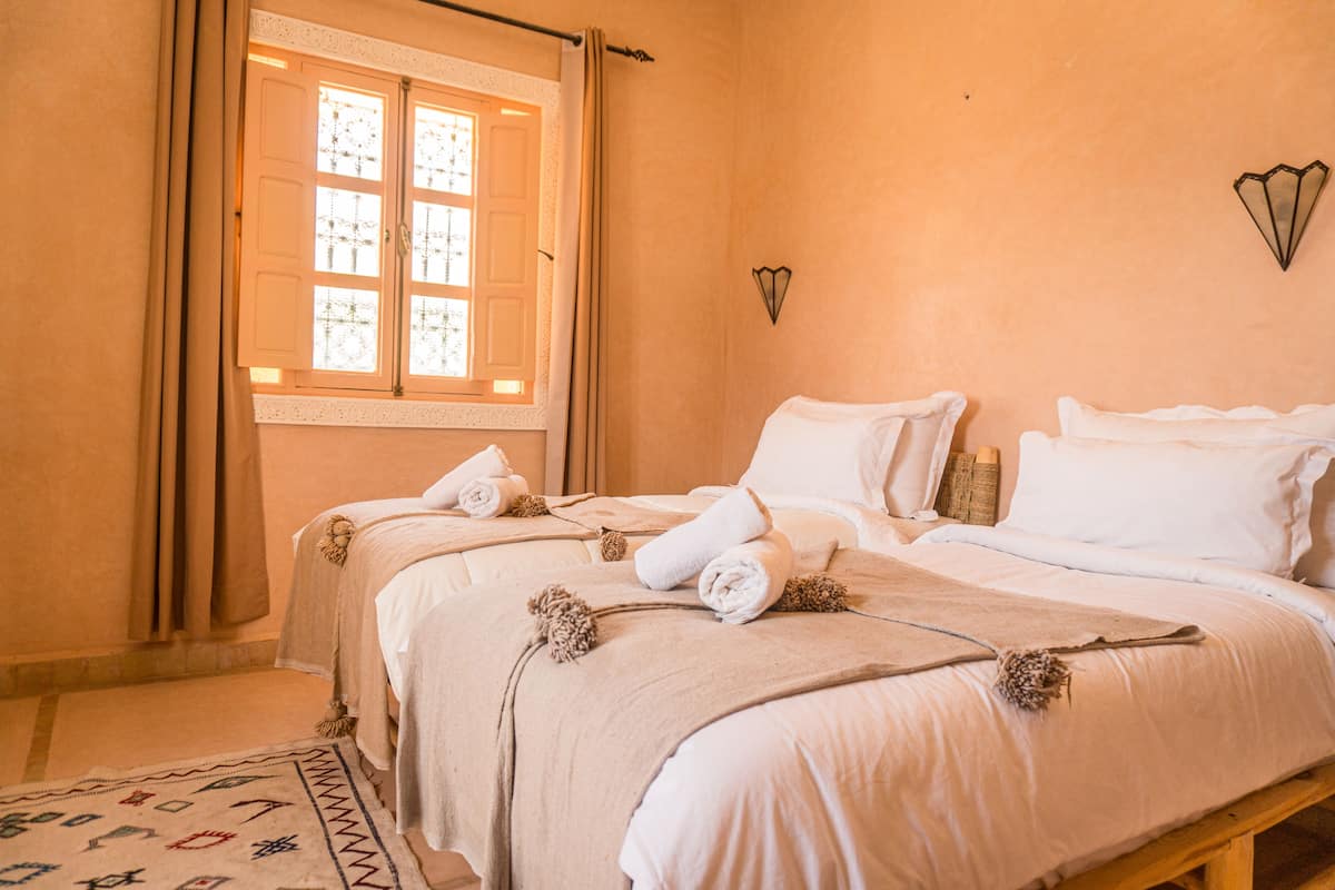 A bedroom with a large bed, a window with curtains, a wall-mounted light fixture, and a patterned rug.