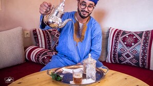 A person in traditional attire pouring tea from a silver teapot onto a tray with a glass and a sugar bowl. - Dar Ajgal (Tanalt)