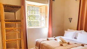 A bedroom with a bed, a wooden ladder, a window with a metal screen, and a wall-mounted light fixture. - Dar Ajgal (Tanalt)