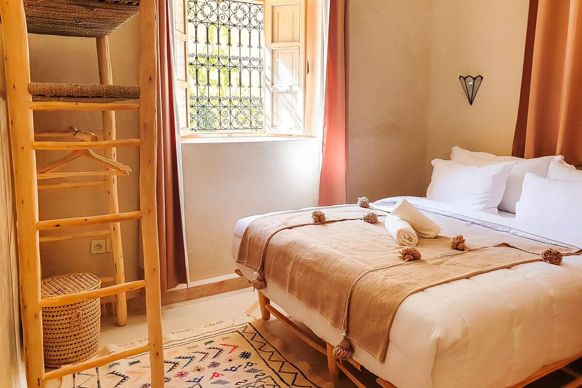 A bedroom with a bed, a wooden ladder, a window with a metal screen, and a wall-mounted light fixture.