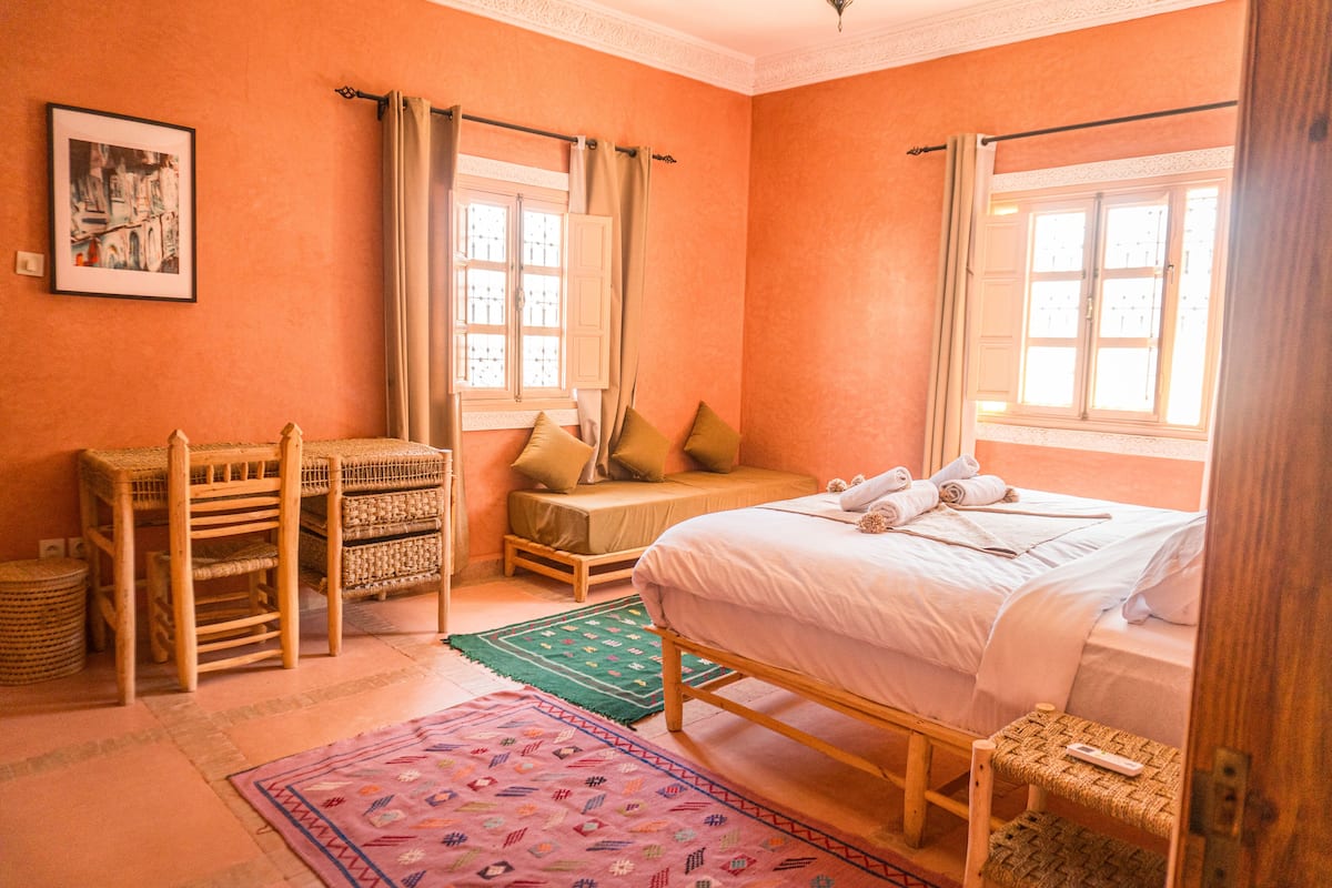 A room with a bed, two windows with curtains, a wicker chair, a small table, and a framed picture on the wall.