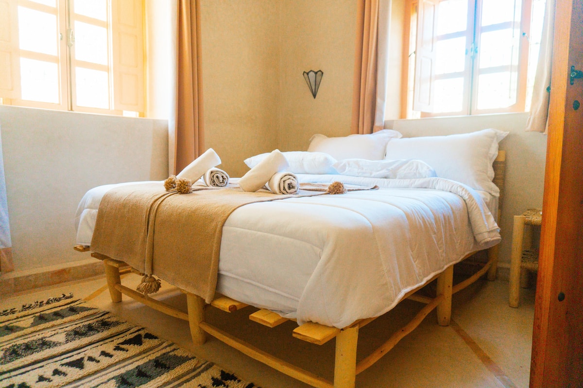 A neatly made bed with white linens and a beige blanket, a wooden headboard, and a small bedside table with a lamp.