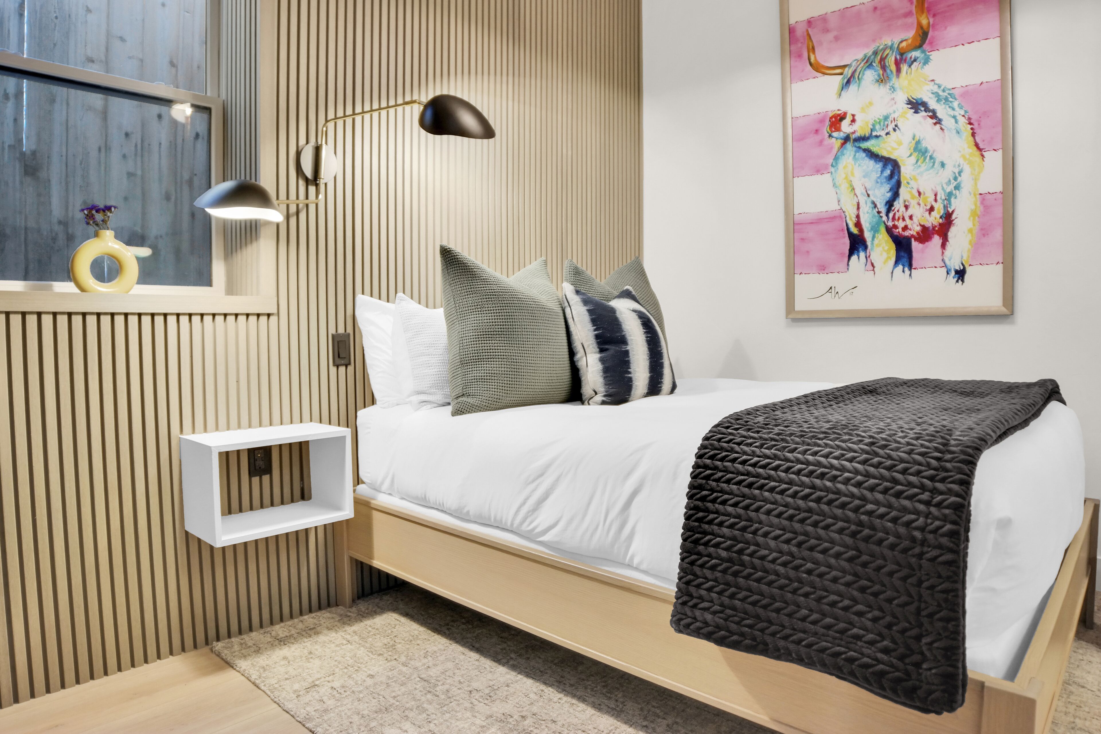 A modern bedroom with a wooden bed, a bedside lamp, a wall-mounted shelf, and a colorful painting.