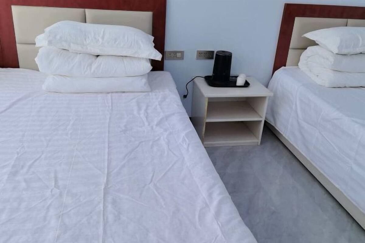 A hotel room with two beds, each with white bedding and pillows, a bedside table with a black object, and a light blue wall.