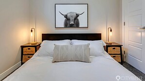 A neatly made bed with a patterned pillow, a framed artwork of a bull's head on the wall, and two bedside tables with lamps. - Blatchford - 2 Queen Beds - W/D to NAIT (Northwest)